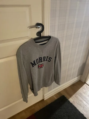 Grå sweatshirt från Morris - Grå sweatshirt från Morris med rund halsringning och tryckt logga samt brittisk flagga på bröstet. Klassisk passform och långa ärmar, perfekt för en avslappnad och stilren look.