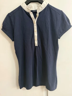 Marinblå kortärmad topp från Gant - Snygg marinblå topp från Gant med vit krage och knappslå. Modellen är kortärmad och har en klassisk, lite preppy look. Tillverkad i mjuk bomull som känns skön mot huden. Perfekt till jeans eller kjol för en clean stil.