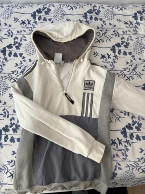 Adidas hoodie med huva och dragkedja - Snygg hoodie från Adidas i beige och grått med huva och halv dragkedja. Tröjan har klassiska Adidas-ränder på axeln, logotyp på bröstet och quiltade grå detaljer på sidorna. Materialet är en mix av bomull och syntet, perfekt för en avslappnad look.    OBS!!! Som ni ser på bilden så finns det fläckar men det går säkert o ta bort.