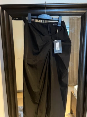 Svart draperad kjol PrettyLittleThing - Snygg svart kjol från PrettyLittleThing med draperad detalj framtill och hög midja. Kjolen har en elegant twistad effekt och stängs med dragkedja bak. Perfekt för dig som vill ha en stilren och trendig look.