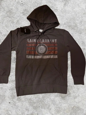 Grå Saint Laurent hoodie  - Snygg grå hoodie från Saint Laurent med huva. Tillverkad i mjukt bomullsmaterial, perfekt för en avslappnad streetwear-look.