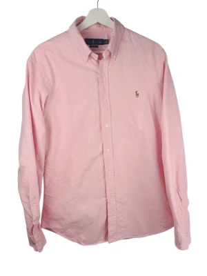 Ralph Lauren Slim Fit Rosa Skjorta, L - Klassisk rosa skjorta från Ralph Lauren med button-down krage och broderad logga på bröstet. Skjortan har lång ärm, knappar framtill och är tillverkad i mjuk bomull. Perfekt för en clean och stilren look.                                      Armhåla till armhåla: 55cm Axelbredd: 46cm Långt: 77cm Armlängden från armhåla: 52cm