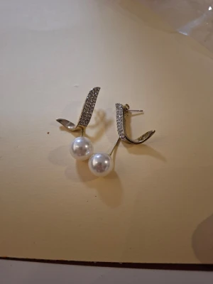 Silvriga örhängen med pärlor - Säljer ett par stilrena örhängen i silverfärg med gnistrande stenar på bågen och vita pärlor som hänger ned. Örhängena har en modern, böjd design och ger en elegant touch till din look.