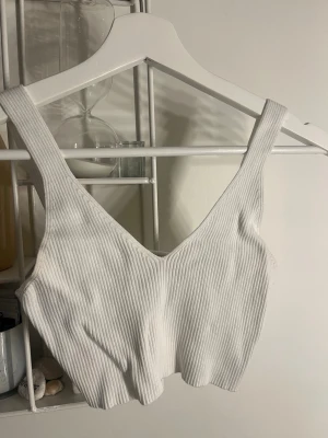 Vit ribbad croppad linnetopp - Säljer en stilren vit croppad linnetopp med breda axelband och v-ringning. Toppen är ribbad och har en mjuk, stretchig känsla som passar perfekt till sommaren. Enkel att matcha med både jeans och kjol för en trendig look.