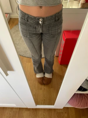 Grå jeans  - Säljer ett par grå lågmidjade bootcut jeans med fyrkantiga fickor framtill. Jeansen har dubbla knappar i midjan och markerade sömmar längs benen för en cool look. 