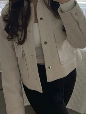 Beige kort cardiganjacka - Stilren beige cardiganjacka med raka linjer och två stora fickor framtill. Jackan har tryckknappar i silver och är tillverkad i ett mjukt, tjockare material som ger en lyxig känsla. Perfekt att slänga över en topp för en clean look.