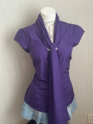 Lila blus med knytband i halsen - Trendig lila blus med korta ärmar och ett långt knytband i halsen som ger en cool och unik look. Blusen har figurnära passform och är tillverkad i ett mjukt, lätt material som känns skönt mot huden. Perfekt för dig som vill sticka ut med färg och stil.