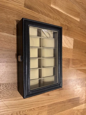 Svart klocklåda med glaslock - Snygg svart klocklåda med plats för 12 klockor. Lådan har ett genomskinligt glaslock och vita sömmar längs kanterna. Insidan är ljusbeige med separata fack och mjuka kuddar för varje klocka. Perfekt för att organisera och visa upp din samling.