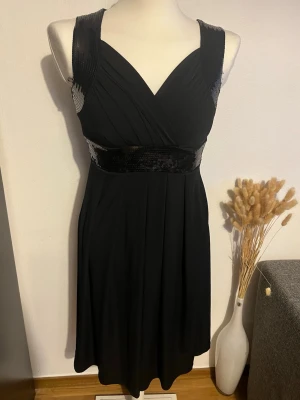 Svart klänning med paljettdetaljer - Snygg svart klänning med breda axelband och v-ringning. Klänningen har glittrande paljetter på axelbanden och i midjan, vilket ger en festlig touch. Materialet är mjukt och faller fint. Perfekt för dig som vill sticka ut med lite extra bling.några paljetter har lossnat 