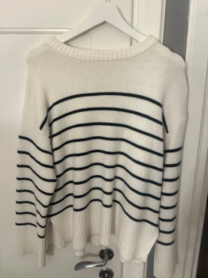 Vit stickad tröja med svarta ränder - Säljer en vit stickad tröja med tunna svarta horisontella ränder. Tröjan har rund halsringning och ribbstickade muddar vid ärmslut och nederkant. Perfekt för dig som gillar klassisk och enkel stil.