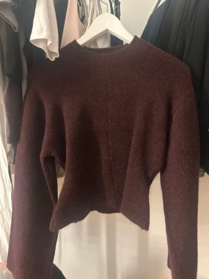 Mörkröd croppad stickad tröja - Mysig mörkröd stickad tröja med croppad passform och långa ärmar. Tröjan har en enkel design med ribbad halsringning och mjukt, fluffigt material som känns skönt mot huden. Perfekt för lager på lager och en chill look.