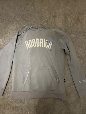 Grå hoodie från Hoodrich - Snygg grå hoodie från Hoodrich med stor logga i vitt på bröstet och mindre tryck på ärmen. Klassisk känguruficka framtill och justerbar huva. Perfekt för en avslappnad streetwear-look.