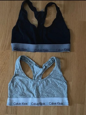 2 Calvin Klein bh-toppar - Säljer två sportiga bh-toppar från Calvin Klein. En är svart med grå resår och logga, den andra är ljusgrå med diskret mönster och vit resår med logga. Båda har brottarrygg och är perfekta för träning eller chill.