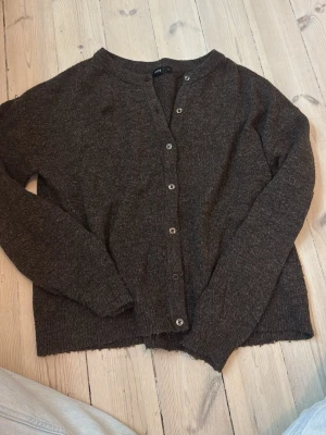 Mörkbrun kofta från LMTD - Mysig mörkbrun kofta från LMTD med rund halsringning och knappar hela vägen fram. Koftan är stickad och har långa ärmar samt ribbade muddar vid ärmslut och nederkant. Perfekt att slänga på sig när det är kyligt ute.