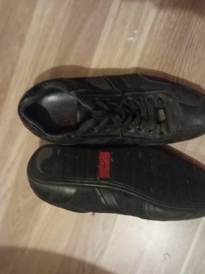 Boss svart skor - Svarta sneakers från boss med klassisk låg profil och snörning. Skorna har diskreta detaljer i svart och en röd boss-logga på sulan. Ovandelen verkar vara i mocka och syntetmaterial, perfekt för en stilren och sportig look.