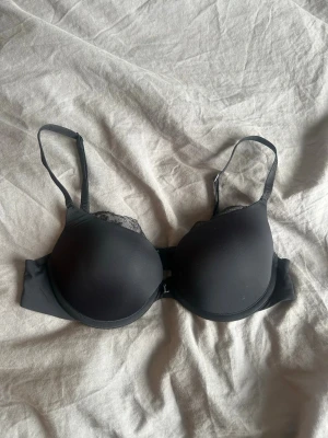Svart push-up bh med spetsdetaljer - Svart push-up bh med vadderade kupor och tunna, justerbara axelband. Bh:n har fina spetsdetaljer vid kupornas övre kant och en slät baksida. Perfekt för dig som vill ha extra lyft och en klassisk look.