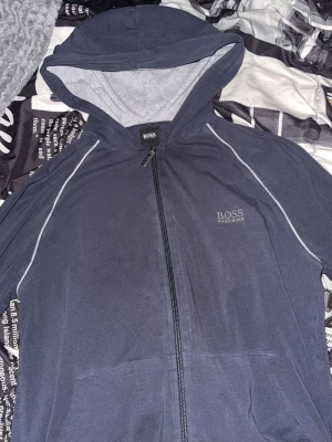 Mörkblå hoodie från Hugo Boss - Snygg mörkblå hoodie från Hugo Boss med dragkedja framtill och gråa detaljer på insidan av huvan och vid muddar. Diskret logga på bröstet och praktiska fickor. Perfekt för en chill och stilren look.