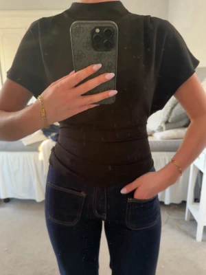Svart topp med hög hals - Säljer en svart topp med hög hals och korta, vida ärmar. Toppen har en figurnära passform i midjan och är tillverkad i ett mjukt material. Perfekt att styla med jeans eller kjol för en clean look.