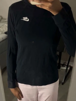 Svart långärmad topp från Nike - Svart långärmad topp från Nike med vit logga på bröstet. Enkel och stilren design med rund halsringning. Perfekt för en sportig och avslappnad look. Dock har tröjan fått en pytte liten färg fläck.