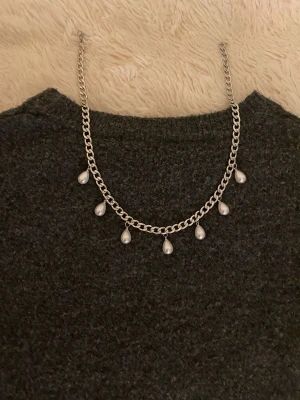 Silverfärgat halsband med pärldroppar - Snyggt halsband i silverfärgad metall med sju droppformade pärlor som hänger längs kedjan. Perfekt för att ge din outfit en elegant touch. Kedjan har en klassisk länkdesign och pärlorna ger ett stilrent intryck.