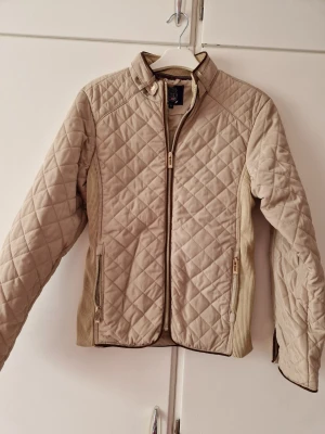 Beige quiltad jacka med dragkedja - Snygg beige quiltad jacka med dragkedja framtill och ståkrage. Jackan har ribbade partier på sidorna och detaljer med knappar vid ärmsluten. Perfekt för dig som gillar stilren och klassisk look.