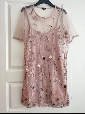 Blommig meshklänning med broderi - Superfin kortklänning från River Island ljusrosa mesh med broderade blommor i olika färger. Klänningen har korta ärmar och en underklänning i matchande färg. Perfekt för dig som gillar romantiska detaljer och transparenta material. Oanvänd.