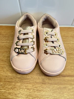 Rosa sneakers från Michael Kors  - Snygga ljusrosa sneakers från Michael Kors med guldfärgade detaljer och logga på snörningen. Skorna har en låg profil, mjuk vadderad insida och guldfärgad häl. Tillverkade i syntetmaterial med klassisk snörning.