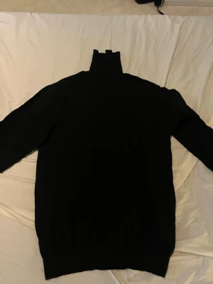 Svart stickad polotröja - En svart stickad polotröja med lång ärm och hög krage. Tröjan har en klassisk rak passform och är perfekt för lager-på-lager-stil. Materialet ser ut att vara mjukt och värmande, perfekt för kyligare dagar.