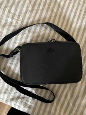 axelväska från Lacoste - Snygg svart axelväska från Lacoste med stilren design och diskret logga framtill. Väskan har justerbar axelrem och dubbla dragkedjor med Lacoste-detaljer. Perfekt för sommaren 
