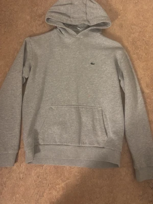 Grå hoodie från Lacoste - Snygg grå hoodie från Lacoste med klassisk krokodillogga på bröstet. Hoodien har huva, magficka och ribbade muddar vid ärmslut och nederkant. Perfekt för en chill och stilren look. den har dock en väldigt väldigt liten fläck precis ovanför fickan men inget som syns jätte mycket 