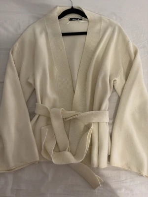 Krämvit cardiganjacka från Gina Tricot - Krämvit cardiganjacka från Gina Tricot med bred ribbad krage och knytskärp i midjan. Jackan har långa ärmar och är tillverkad i ett mjukt stickat material. Perfekt för lager-på-lager och en chill look. Man kan även knyta bandet baktill för en mer kroppad look.