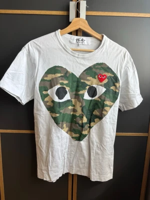 Vit Comme des Garçons Play t-shirt - Vit t-shirt från Comme des Garçons Play med stort hjärta i camouflage-mönster och ögon på bröstet. Liten röd hjärtpatch med ögon på vänster sida. Klassisk rund hals och korta ärmar. Perfekt statement-plagg för din garderob.