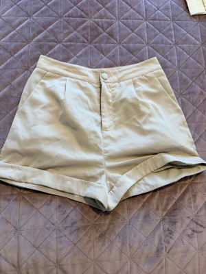 Beiga höga shorts med fickor - Stilrena beiga shorts med hög midja och uppvikta benslut. De har två sidofickor och stängs med knapp och dragkedja framtill. Perfekta för varma dagar och enkla att matcha med olika toppar.