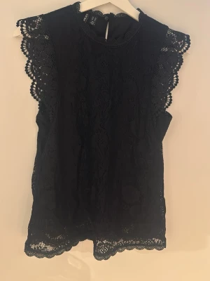 Svart spetsblus från Pieces - Supersnygg svart blus från Pieces med spetsdetaljer och vackert mönstrade kanter. Blusen är ärmlös med rund halsringning och knäppning bak i nacken. Perfekt att styla med jeans eller kjol för en trendig look.