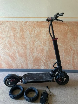 Svart elscooter med extra däck - Säljer en svart elscooter med robust ram och bred ståplatta. Medföljer två extra däck och laddare. Scootern har röda detaljer vid hjulupphängningen och handtaget. Perfekt för dig som vill ta dig fram snabbt och smidigt i stan.