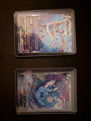 Pokémonkort Vaporeon VMAX & V - Två samlarkort från Pokémon med Vaporeon VMAX och Vaporeon V. Båda korten har färgstarka illustrationer och ligger i genomskinliga plastfickor. Perfekt för dig som gillar Pokémon och vill utöka din samling med snygga V och VMAX-kort.