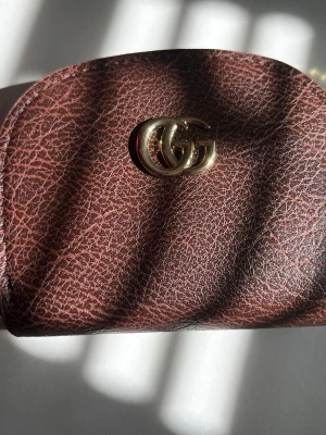 Brun plånbok från GG - Säljer en stilren brun plånbok med GG märke, klassisk rundad form och snygg läderstruktur. Perfekt för dig som gillar en tidlös och enkel design. Plånboken har en mörkbrun färg och är gjord i material som liknar läder.
