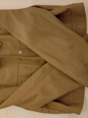 Beige skinnjacka från NA-KD - Stilren beige skinnjacka från NA-KD med klassisk krage, tryckknappar och två stora bröstfickor. Jackan har raka linjer och en modern, boxig passform som ger en cool vibe till din outfit.