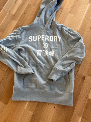 Superdry hoodie Storlek S Herr - Snygg grå hoodie från Superdry med vit logga och japanska tecken på ryggen. Hoodien har huva, magficka och ribbade muddar. Perfekt för en avslappnad streetstyle-look.