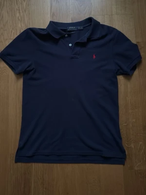 Marinblå pikétröja från Polo Ralph Lauren - Klassisk marinblå pikétröja från Polo Ralph Lauren med korta ärmar och krage. Tröjan har två knappar vid halsen och en liten röd broderad logga på bröstet. Perfekt till jeans eller shorts.