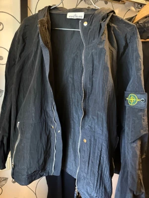 Svart vindjacka från Stone Island - Svart vindjacka från Stone Island med huva och dragkedja framtill. Jackan har två fickor med dragkedja och den klassiska logotypen på vänster ärm. Lättviktigt material som ger en sportig och stilren look.