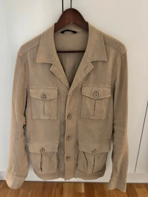 Beige safarijacka med fickor, Oscar Jacobsson - Stilren beige safarijacka från Oscar Jacobsson med fyra stora fickor framtill och knäppning med knappar. Jackan har klassisk krage och är tillverkad i ett mjukt bomullsmaterial. Perfekt för en avslappnad och trendig look.