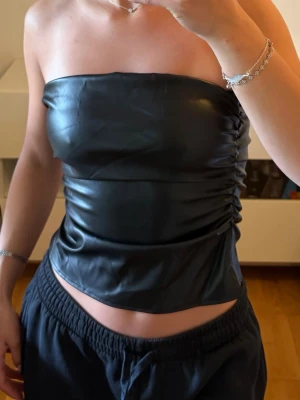 Svart tubtopp i skinnimitation - Säljer en svart axelbandslös tubtopp i skinnimitation med snygg rynkning på sidan. Toppen har en glansig finish och sitter tight över bröstet. Perfekt för dig som vill ha en edgy och trendig look.