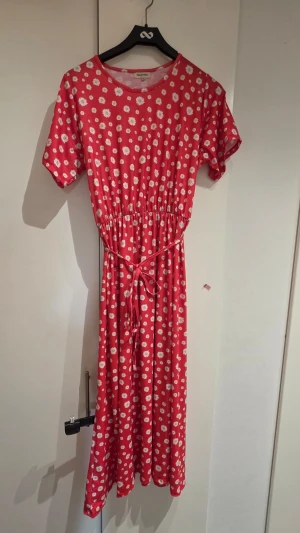 Röd blommig klänning från Hängmatta - Söt röd långklänning från Hängmatta med vita och gula blommor, korta ärmar och knytband i midjan. Klänningen har rund halsringning och är tillverkad i mjuk bomull, perfekt för varma dagar.