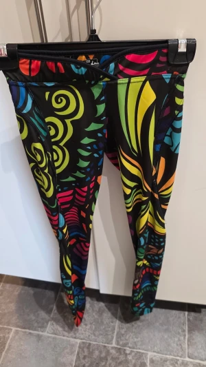 Färgglada mönstrade leggings XS - Säljer ett par leggings med färgstarkt och psykedeliskt mönster i neonfärger som gult, grönt, blått, rött och orange på svart botten. De är stretchiga och sitter tight, perfekta för dig som vill sticka ut. Materialet känns som polyester eller liknande.