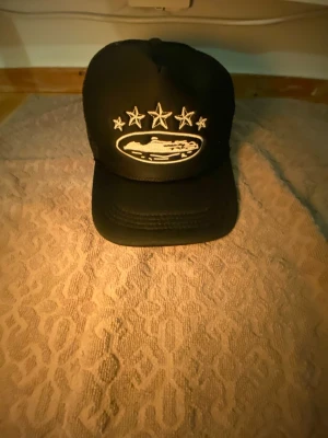 Svart truckerkeps med broderade stjärnor - Svart truckerkeps med vit brodyr framtill och på sidan. Framsidan har ett broderat motiv med fyra stjärnor och ett ovalt emblem. Baksidan är i mesh för extra ventilation. Perfekt för en cool och avslappnad stil.