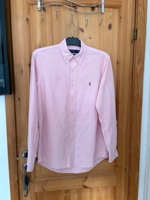 Ljusrosa skjorta från Polo Ralph Lauren - Klassisk ljusrosa skjorta från Polo Ralph Lauren med button-down krage och broderad logga på bröstet. Skjortan har lång ärm, knäppning framtill och är tillverkad i mjuk bomull. Perfekt för en clean och stilren look.