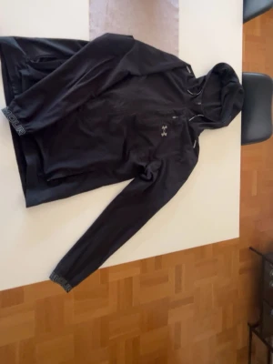 Svart hoodie från Under Armour - Säljer en svart sportig hoodie från Under Armour med dragkedja och huva. Tröjan har långa ärmar och logga på bröstet samt textdetaljer på ärmslutet. Perfekt för träning eller chill. Materialet är mjukt och stretchigt för extra komfort.