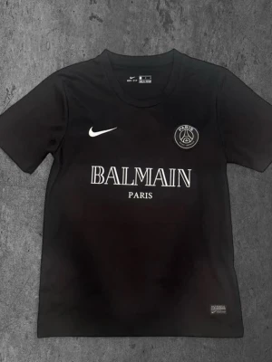 Svart PSG x Balmain t-shirt Nike - Säljer en svart t-shirt från Nike med Paris Saint-Germain x Balmain Paris tryck på bröstet. Tröjan har rund hals, korta ärmar och är tillverkad i Dri-FIT material som håller dig torr. Perfekt för dig som gillar exklusiva samarbeten och sportmode.