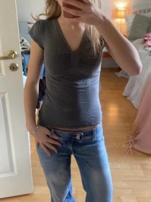 V-ringad topp - Snygg mörkgrå topp med v-ringning och rynkdetalj framtill. Modellen är figurnära med korta ärmar och mjukt, stretchigt material. Perfekt för en trendig och avslappnad look.